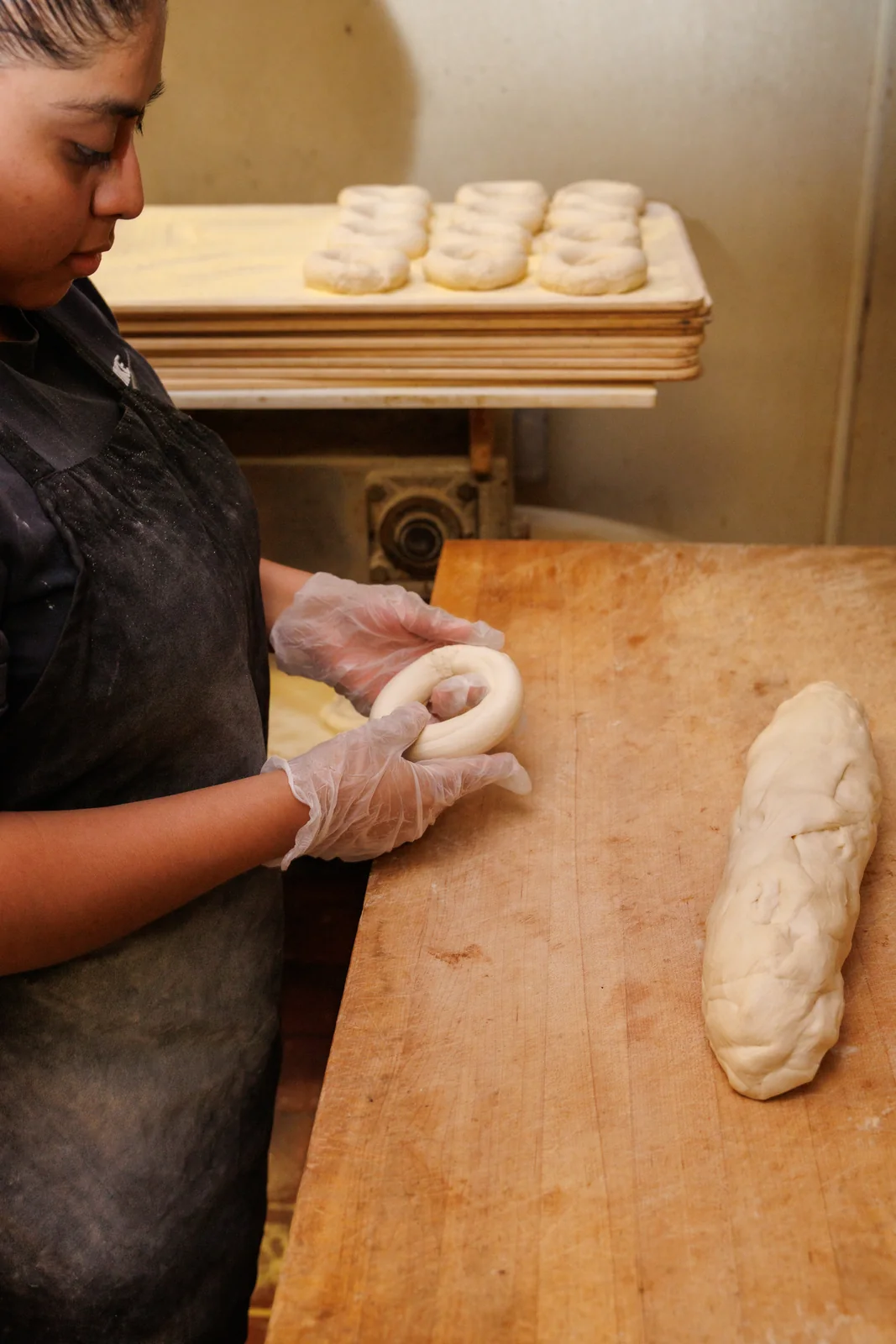 A Bagel Boyz baker shaping dough by hand, with trays of bagels behind the bench